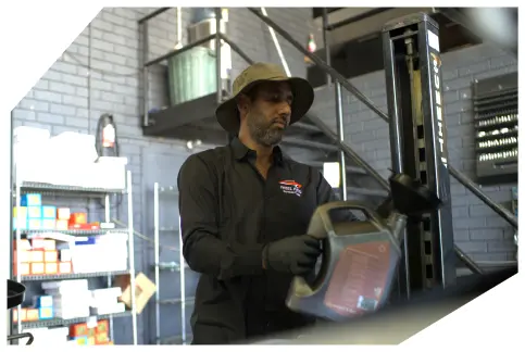Mask-group A Patel Auto Services worker in a hat pours oil in a garage with tools and shelves nearby.