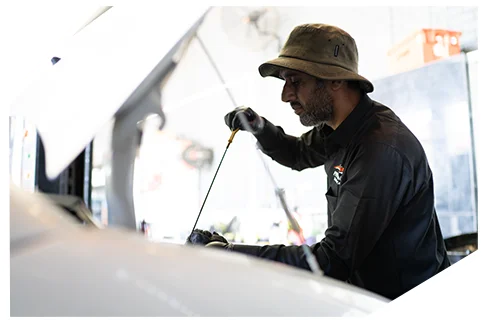 Basic Oil Service03 A mechanic wearing a bucket hat and dark uniform checks oil levels using a dipstick while working under an open vehicle hood in a repair garage.