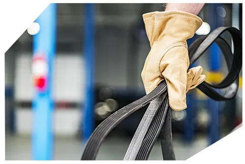 Belts & Hoses04 Tan leather work glove holding black serpentine belts in an auto repair shop, with blurred blue service equipment visible in the background.