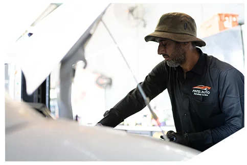 Electrical & Wiring02 Auto mechanic wearing a bucket hat and dark shirt with patel auto services logo working under an open car hood in a repair garage.