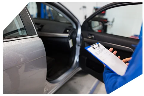 Electrical & Wiring04 Technician in blue uniform holding clipboard while inspecting car interior through open driver's door in auto service garage.