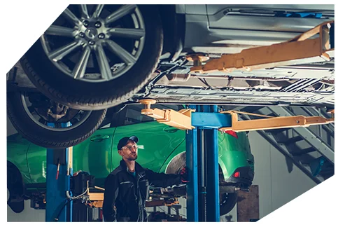 Genaral Automotive 05 A mechanic in a black uniform and cap inspects a car lifted on a hydraulic lift in a well-equipped auto repair workshop with multiple vehicles.