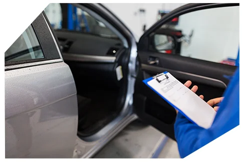 Pink Slip & Registration002 A mechanic in a blue uniform is holding a clipboard and assessing a silver car with its door open. The person is likely performing a vehicle inspection details about the car’s condition in a professional workshop.