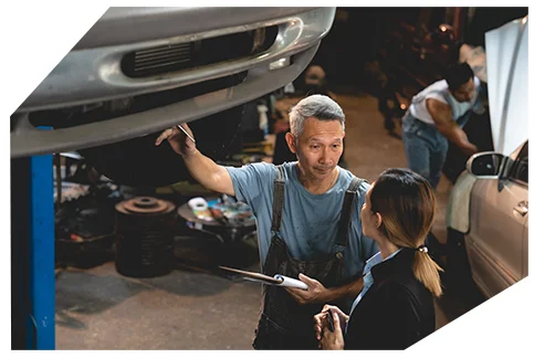 Pink Slip & Registration003 A mechanic in overalls explains a car issue to a woman in a suit, pointing at a lifted vehicle in a busy auto shop.