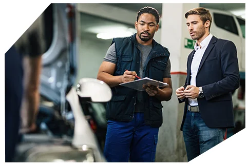 Pink Slip & Registration004 A mechanic in a vest reviews a clipboard with a man in a suit inside an auto shop, discussing car repairs near a vehicle.