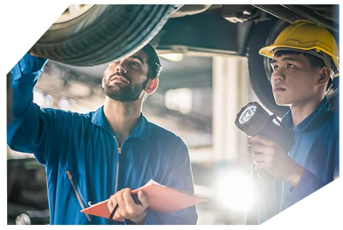 Pink Slip & Registration006 Two mechanics in blue uniforms, one with a hard hat and flashlight, inspect a car's undercarriage on a lift, holding a clipboard.