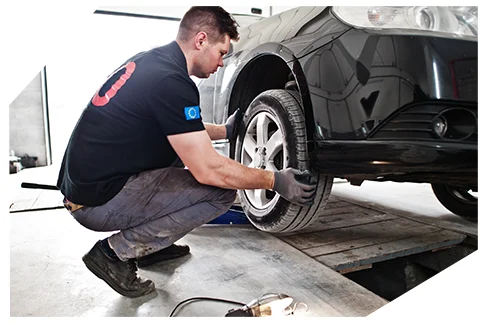 Safety & Tyre Checks01 A mechanic inspecting or working on a car's front wheel. The car is lifted slightly on a platform, and the mechanic is crouching while carefully checking the tyre.