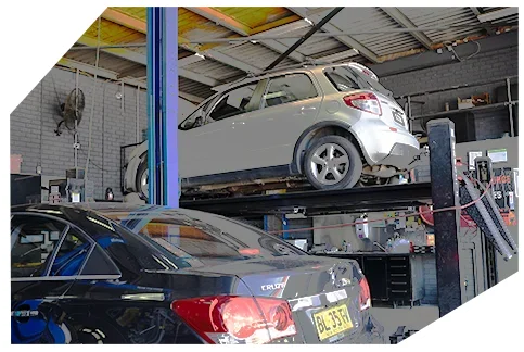 Suspension & Steering A car repair workshop with a silver hatchback elevated on a hydraulic lift and a black sedan parked below, surrounded by tools and equipment.