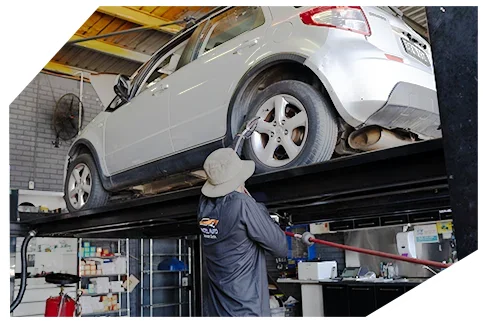 Suspension & Steering02 A mechanic wearing a hat and dark uniform uses a power tool to work on a silver car lifted on a hydraulic platform in a workshop.