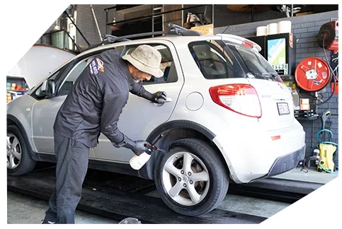 Suspension & Steering03 A mechanic wearing a hat and gloves cleans a silver car’s wheel and body using a spray bottle while working in a well-equipped auto repair workshop.