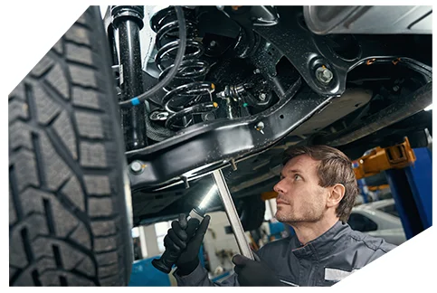 Suspension & Steering04 A mechanic in gloves and a gray uniform inspects a car's undercarriage using a flashlight and tool while the vehicle is lifted on a hydraulic lift.