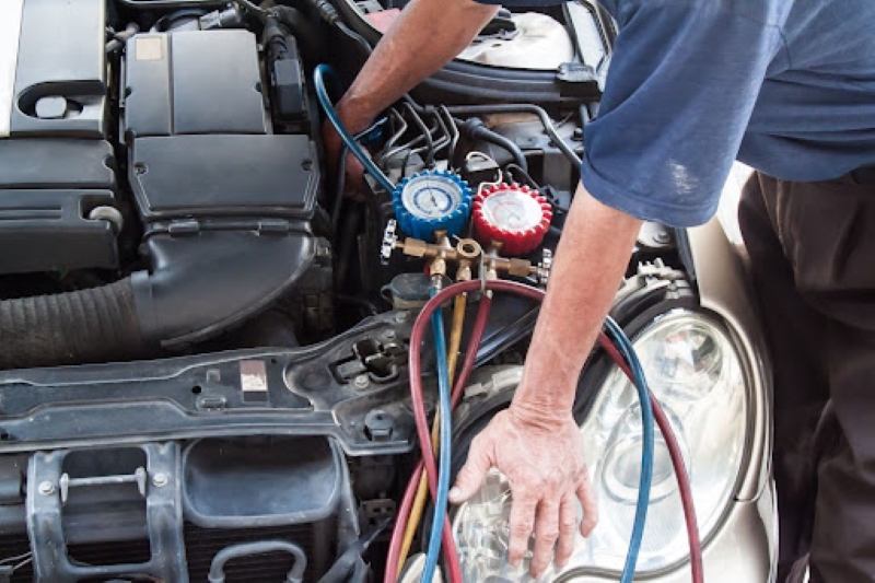 A person working on a car’s air conditioning system under the hood, using gauges and hoses connected to the engine in a garage setting.