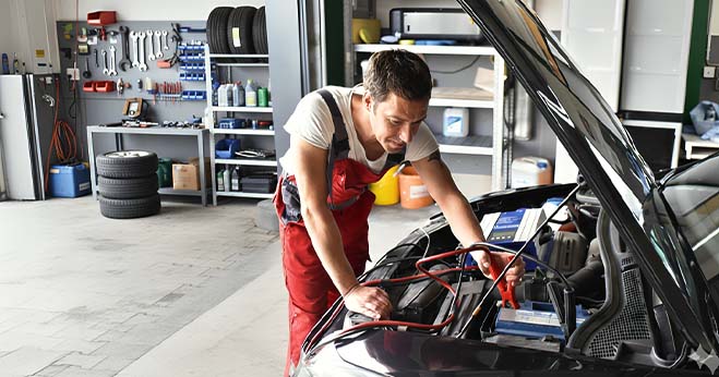 Mechanic in blue overalls checking off items on a 'Vehicle Maintenance Checklist' clipboard in front of an open car engine.