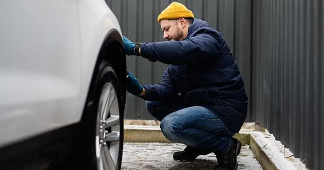 A man in a yellow beanie and blue jacket performing a manual tyre tread and pressure check on a white car.