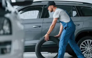 Mechanic in blue overalls carrying a spare car tyre in a garage setting.
