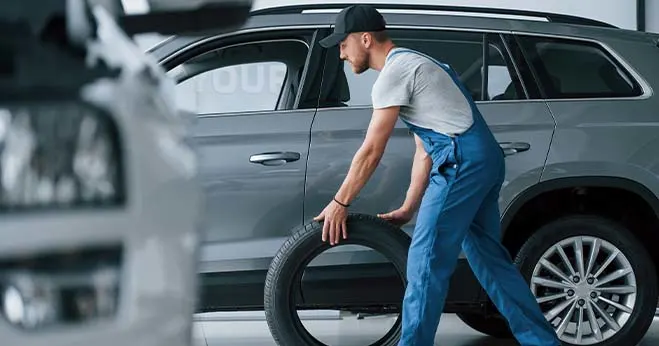 Mechanic in blue overalls carrying a spare car tyre in a garage setting.